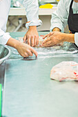 Crop anonymous cooks in uniforms and aprons rolling raw meat in clingfilm while cooking together