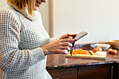 Crop smiling adult lady in cozy sweater tapping screen of tablet while standing against cabinets in kitchen at home