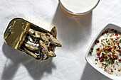 Draufsicht auf Sardellen in Dosen, serviert auf einem weißen Tisch mit einem Glas schäumendem Bier und weißer Gazpacho-Suppe