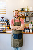 Positive male barista in apron looking at camera with crossed arms while standing near counter with paper cups and desserts in coffee house