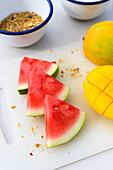 Top view of fresh sliced watermelon and orange juicy cut in half mango placed on white chopping board with knife