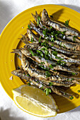 Top view plate of appetizing grilled anchovies served on table with piece of lemon and fresh herbs in restaurant