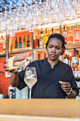 Focused African American female barkeeper pouring alcohol in glass with ice cube while making cocktail in bar