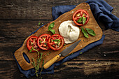 Top view of delicious mozzarella and slices of fresh tomato with green basil leaves and thyme served on wooden cutting board with knife on table