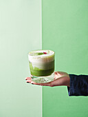 Anonymous person demonstrating glass of delicious refreshing matcha latte with whipped milk on green background