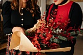 Cropped unrecognizable female friends standing at table with candles and making creative Christmas bouquets for holiday celebration