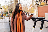 Hispanische langhaarige Frau in warmem Mantel steht auf einer Straße in der Stadt und bezahlt kontaktlos einen Elektroroller mit einem Smartphone über eine Anwendung