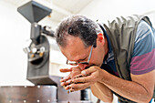Side view of male employee in eyeglasses pouring coffee beans from hands and inhaling aroma in light room
