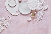 Draufsicht auf einen Satz verschiedener weißer Keramikteller mit Schneidebrett und kleinem Krug auf einer blassrosa Fläche mit Stoff