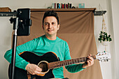 Cheerful adult male vlogger with guitar and plectrum recording video tutorial on cellphone at home