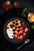 Top view of yummy healthy homemade breakfast with fresh assorted berries served with yogurt and cookies in black plate on table with ingredients