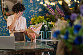 Cheerful female florist speaking on mobile phone and getting order while standing in floral store with flower bouquet