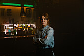 Attractive waitress in apron looking at camera with crossed arms while standing near bar counter with bottles of alcohol drinks