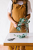 Crop unrecognizable female florist in apron creating composition with fresh green Eucalyptus leaves standing at table in light studio