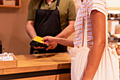 Unrecognizable woman with tote bag using credit card to pay for goods to vendor in zero waste grocery store