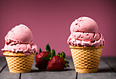 Yummy pink ice cream scoops with waffle cones placed on table near red ripe strawberries
