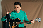 Cheerful adult male vlogger with guitar and plectrum recording video tutorial on cellphone at home