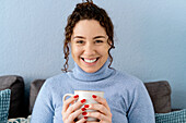 Calm female with curly hair drinking hot beverage from white cup while sitting looking at camera smiling on couch at wall in light room