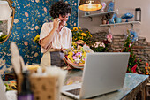 Cheerful female florist speaking on mobile phone and getting order while standing in floral store with flower bouquet