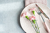 Von oben gedeckter Tisch mit Teller und Serviette neben rosa Nelkenblüten auf Betontisch