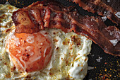 From above of sunny side up egg with fried bacon slices and condiments on dark tray