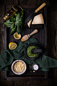 Komposition von oben mit Zutaten für eine traditionelle Pesto-Soße, darunter Parmesankäse, Basilikumblätter, Zitrone und Pinienkerne, auf einem dunklen Tisch