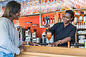 Cheerful black female barkeeper serving glass of refreshing cocktail for client in bar