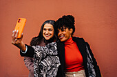 Mature glad diverse female friends taking selfie on phone with tooth smiles while standing near red wall
