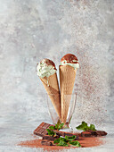 Delicious ice cream scoops in waffle cones with fresh mint leaves and chocolate placed in glass vase against gray background
