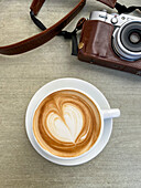 Draufsicht auf einen Becher mit heißem Kaffee mit gezeichnetem Herz, der auf einem Tisch neben einer alten Fotokamera in einer braunen Ledertasche steht