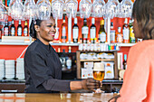 Cheerful black female barkeeper serving glass of refreshing beer for client in bar