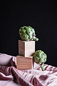 Ripe artichokes placed on wooden cube on table with silk fabric against black background