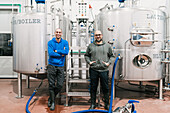 Content male coworkers looking at camera while standing between metal tanks in brewing company