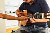 Ausschnitt nicht erkennbarer Musiker, der auf einen Bund zeigt, während er einer anonymen männlichen Person das Spielen einer akustischen Gitarre in einem hellen Raum am Tag beibringt