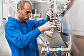 Side view of male worker in sterile mask pouring fermented beer from metal vessel into glass in factory