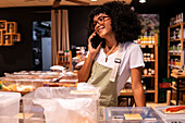 Glad young African American woman in apron and glasses leaning on counter and talking on cellphone while working in zero waste grocery store