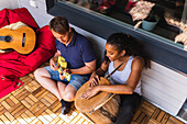 From above of cheerful young diverse couple resting and playing ukulele and djembe sitting on balcony in sunlight