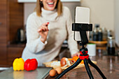 Soft focus on smartphone in tripod placed on stone counter with blurred female vlogger sharing advice on cooking vegetables and smiling