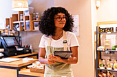 Young black female vendor in uniform and glasses with tablet smiling and inspecting pasta in glass container during work in zero waste grocery store looking away