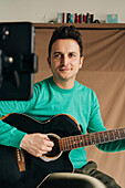 Crop cheerful adult male vlogger with guitar and plectrum recording video tutorial on cellphone at home