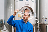 Cheerful male entrepreneur with alcoholic liquid in flask working against fermentation tanks in brewery