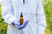 Crop anonymous female scientist wearing white robe and latex gloves demonstrating glass bottle with liquid drug