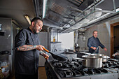 Professional chef doing flambe on food in frying pan with fire while standing near cooker in light kitchen of restaurant