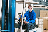 Male engineer in sterile mask driving vehicle and looking forward against cardboard boxes and bottles with beer in factory