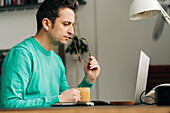 Side view of male distance workers with mug of hot drink against netbook in house