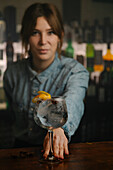 Soft focus female bartender looking at camera while serving wineglass with gin and tonic cocktail decorated with orange zest on wooden counter in bar