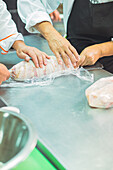 Crop anonymous cooks in uniforms and aprons rolling raw meat in clingfilm while cooking together