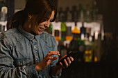 Positive female barkeeper surfing internet on cellphone while standing in bar with various alcoholic drinks during work in modern cafe