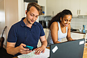 Smiling young multiracial couple using gadgets and devices while sitting at counter in kitchen and studying