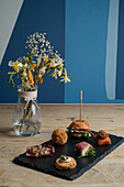 Assorted delicious appetizers served on black board near glass vase with blooming bouquet placed on wooden surface